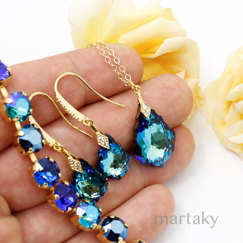 Puede incluir: Un conjunto de collar y pendientes dorados con cristales azul y verde iridiscentes en forma de l&aacute;grima. Los pendientes tienen un dise&ntilde;o de gancho con peque&ntilde;os cristales transparentes. Tambi&eacute;n se ve una pulsera a juego con cristales azules redondos.