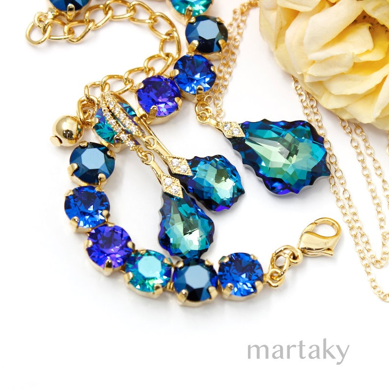 Puede incluir: Un conjunto de joyas doradas con pulsera, collar y pendientes. El conjunto incluye piedras de cristal azules y turquesas. Los pendientes y el collar tienen colgantes en forma de l&aacute;grima. La pulsera tiene piedras redondas. Las joyas est&aacute;n sobre una superficie blanca.