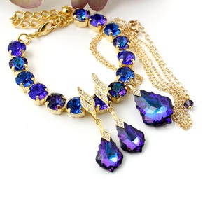Pode incluir: Um bracelete de corrente dourada com uma fila de cristais iridescentes azuis e roxos. O bracelete tem tr&ecirc;s cristais pendentes em forma de l&aacute;grima.