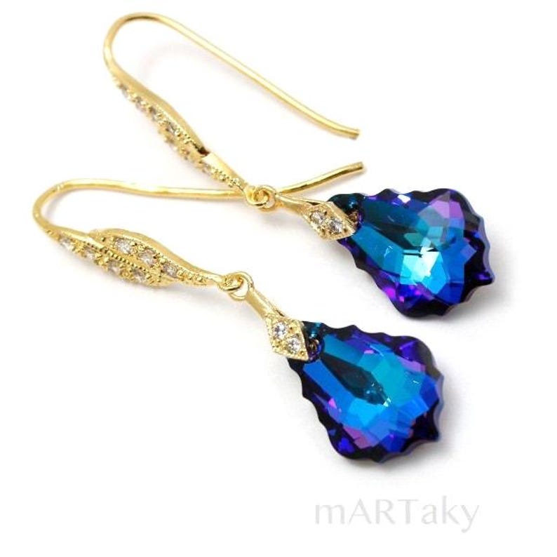 Pode incluir: Brincos de argola dourados com um cristal azul em forma de l&aacute;grima. O cristal tem um acabamento iridescente e est&aacute; montado em um fio dourado.