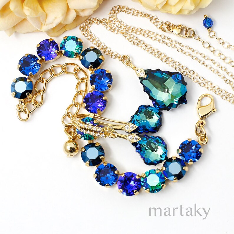 Puede incluir: Conjunto de joyas doradas con detalles de cristales azules y turquesas. El conjunto incluye un collar con colgante de l&aacute;grima, una pulsera y pendientes. Las joyas est&aacute;n dispuestas sobre una superficie blanca.