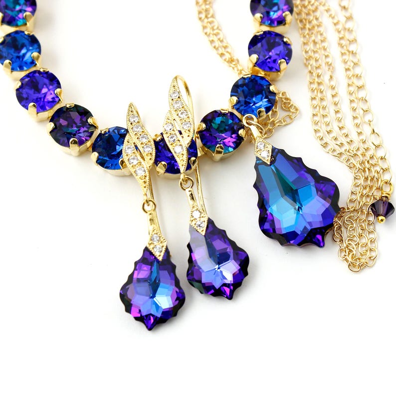 Pode incluir: Um conjunto de colar e brincos de ouro com pedras de cristal iridescentes azuis e roxas. O colar tem uma corrente de v&aacute;rias voltas e os brincos t&ecirc;m forma de l&aacute;grima.