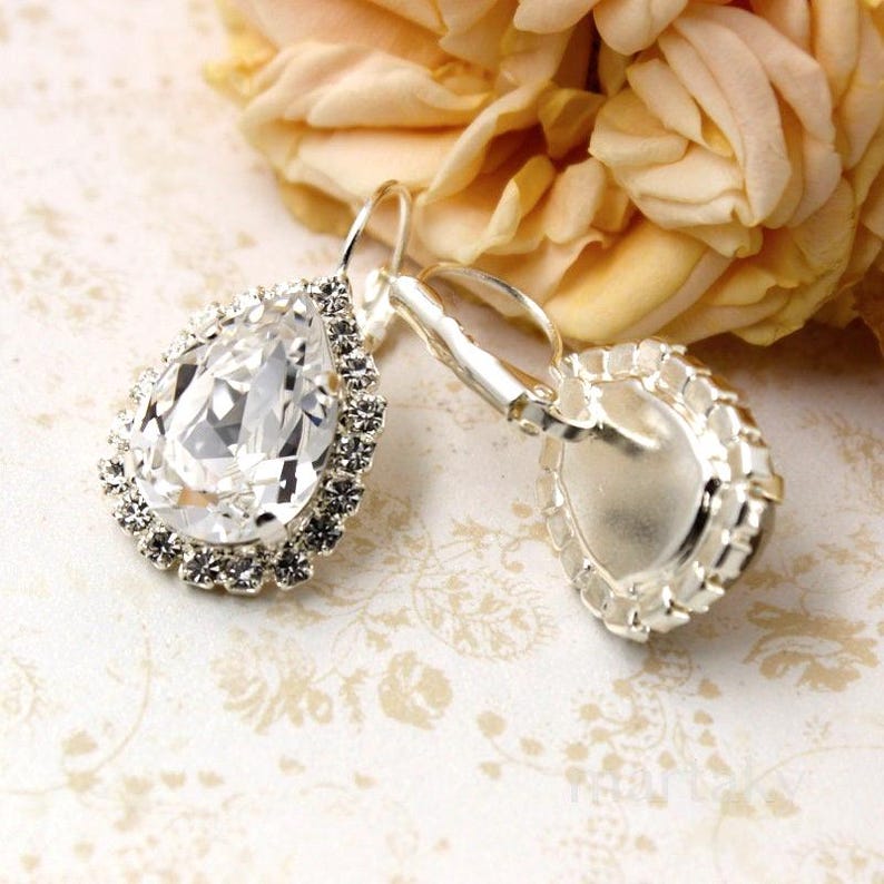 May include: A pair of silver-toned drop earrings. Each earring features a large, clear, teardrop-shaped crystal surrounded by smaller, clear crystals. The earrings are set against a soft, floral background.