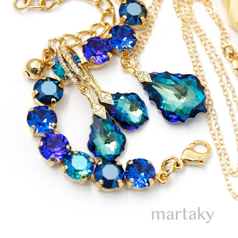 Puede incluir: Un conjunto de joyas dorado que incluye una pulsera, aretes y un collar. El conjunto incluye piedras de cristal azules y turquesas. La pulsera y los aretes tienen un dise&ntilde;o de l&aacute;grima. El collar tiene un colgante grande en forma de l&aacute;grima. La marca "martaky" es visible.