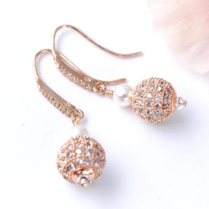 May include: A pair of rose gold earrings with a delicate design. Each earring features a small, round, pearl-like bead and a larger, sparkling, crystal-covered ball. The earring hooks are adorned with small crystals.