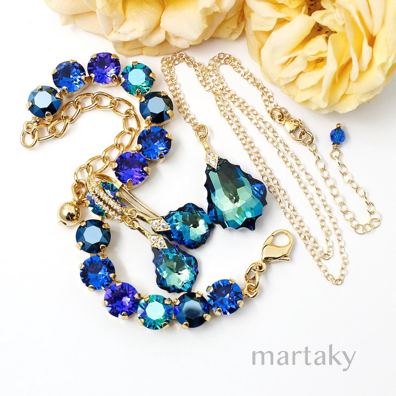 Puede incluir: Un conjunto de joyas en tono dorado con pulsera, collar y pendientes. La pulsera y los pendientes tienen detalles de cristal azul y turquesa. El collar tiene un colgante grande en forma de l&aacute;grima. Las joyas se exhiben sobre una superficie blanca.
