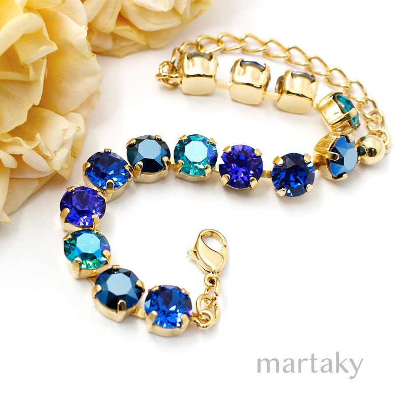 Puede incluir: Una pulsera dorada con una serie de piedras preciosas redondas, facetadas, azules y turquesas. La pulsera tiene una cadena y un cierre de broche. Las piedras est&aacute;n engastadas en metal dorado. Una flor amarilla est&aacute; en el fondo.