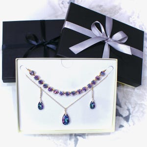 Puede incluir: Un conjunto de joyas con un collar y pendientes con cristales iridiscentes morados y azules. El conjunto se exhibe en una caja blanca, con dos cajas de regalo negras con cintas en el fondo. El collar y los pendientes tienen una cadena dorada.