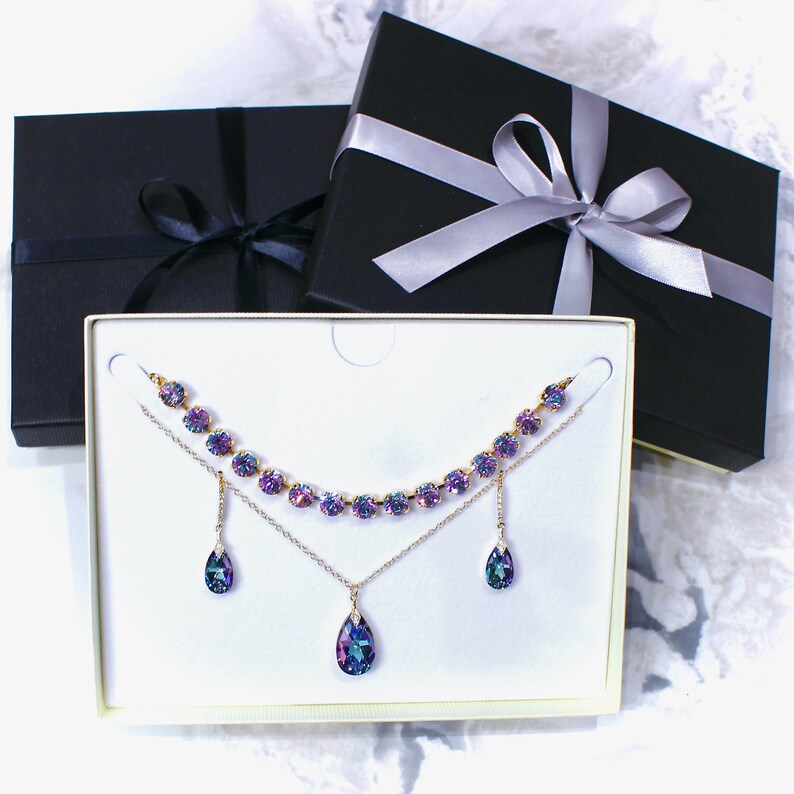 Puede incluir: Un conjunto de joyas con un collar y pendientes con cristales iridiscentes morados y azules. El conjunto se exhibe en una caja blanca, con dos cajas de regalo negras con cintas en el fondo. El collar y los pendientes tienen una cadena dorada.