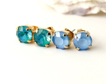 May include: Two pairs of stud earrings. One pair features teal-coloured, faceted stones set in gold-coloured metal. The other pair has light blue stones in a similar setting. The earrings are displayed on a white surface.