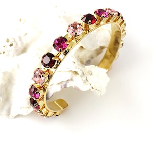 Può includere: Un bracciale a polsino dorato ornato da una fila di pietre preziose rotonde e sfaccettate in tonalità rosa e rosse. Il bracciale è posto su uno sfondo bianco strutturato, che mette in risalto il design e la lucentezza del gioiello.