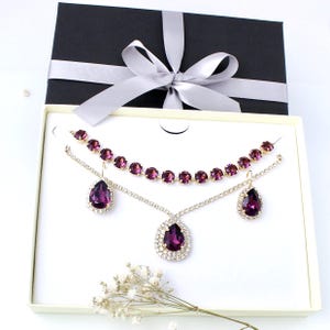 May include: A jewelry set featuring a necklace, earrings, and a pendant, presented in a gift box. The set includes a necklace with round, purple gemstones and a pendant with a large, teardrop-shaped purple gemstone, surrounded by smaller clear stones.