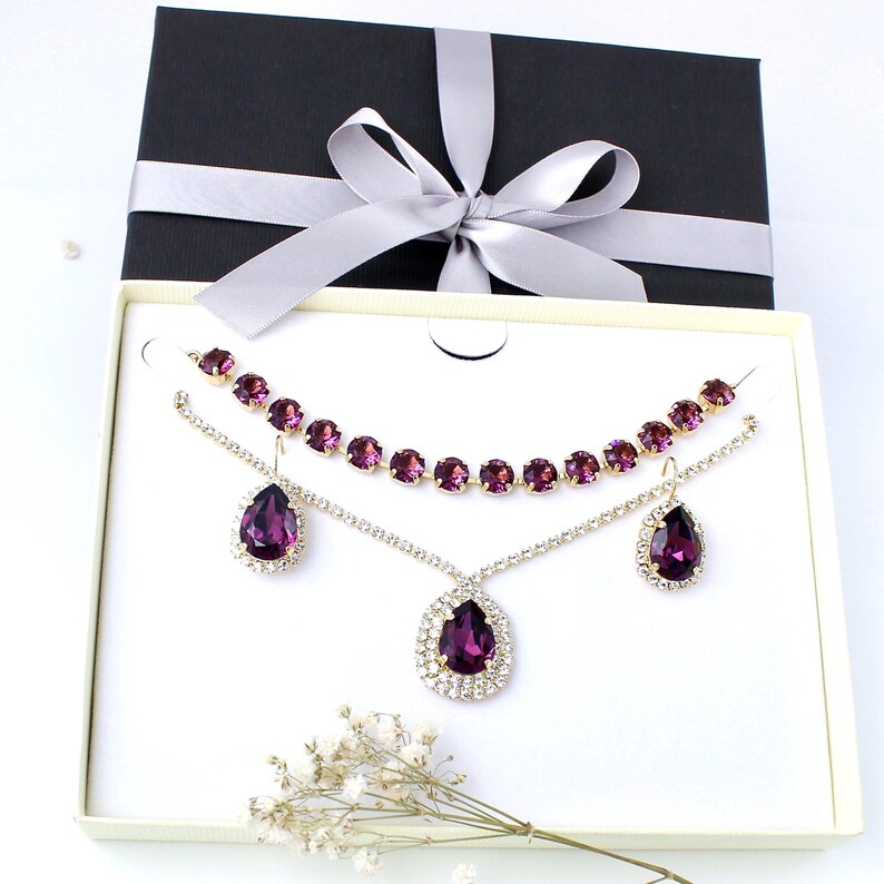 May include: A jewelry set featuring a necklace, earrings, and a pendant, presented in a gift box. The set includes a necklace with round, purple gemstones and a pendant with a large, teardrop-shaped purple gemstone, surrounded by smaller clear stones.