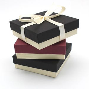 May include: Three square gift boxes stacked on top of each other. The top box is black with a white ribbon, the middle box is burgundy, and the bottom box is black.