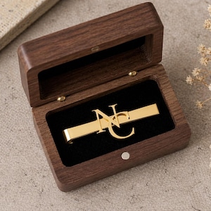 May include: A gold-colored tie clip with the initials "NC" is displayed inside an open, dark wooden box lined with black velvet. The box is rectangular with a hinged lid and brass-colored hardware.