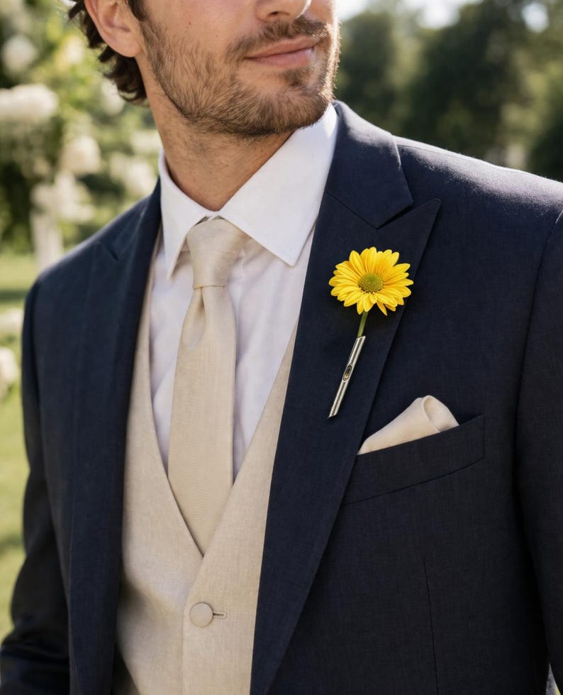 May include: A navy blue suit with a white dress shirt, beige vest, and tie. A yellow flower is in the lapel pocket. A beige pocket square is in the suit's breast pocket. The suit is tailored and appears to be made of a fine fabric.
