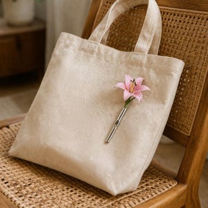 May include: A beige tote bag with a pink lily in a silver bud vase rests on a wooden chair with a woven seat. The bag has two handles and appears to be made of a soft, textured fabric. The chair has a light brown finish.