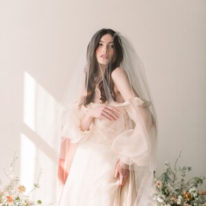 Könnte beinhalten: Eine Braut in einem weißen, schulterfreien Kleid mit langem Schleier. Das Kleid hat einen Rüschen-Oberteil und der Schleier ist durchsichtig und zart. Die Braut steht in einem Raum mit weißem Hintergrund und natürlichem Licht.