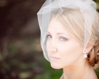 Peut inclure: Une femme blonde, légèrement maquillée, porte un voile blanc transparent. Elle porte également des boucles d'oreilles pendantes en perles. L'arrière-plan est flou, avec une lumière naturelle douce.