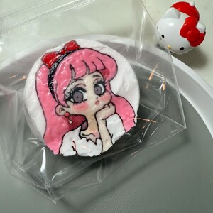 May include: A pink and white cake decorated with a hand-painted image of a girl with pink hair and a red bow. The cake is in a clear plastic bag.