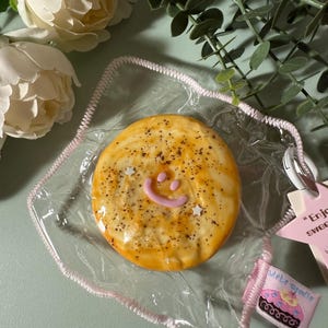 Peut inclure: Un biscuit jaune et brun avec un smiley rose et des étoiles blanches sur le dessus. Le biscuit est dans un sac en plastique transparent avec une bordure rose. Le sac a une étiquette rose et blanche qui dit "WeIrD sTuDiO" avec un graphique de cupcake.