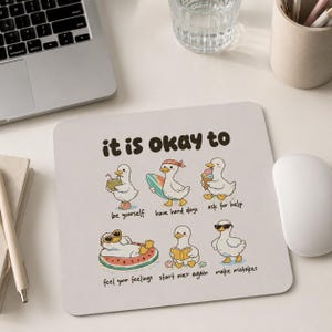 May include: A square mouse pad with a light background featuring six cartoon ducks and the text "it is okay to". The ducks are shown with captions: "be yourself", "have hard days", "ask for help", "feel your feelings", "start over again", and "make mistakes".
