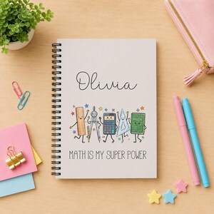 May include: A spiral-bound notebook with a white cover featuring the name Olivia in black script, followed by a colorful illustration of cartoon math supplies and the text MATH IS MY SUPER POWER in black capital letters at the bottom.