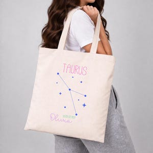 May include: A beige canvas tote bag featuring the word TAURUS in pink, a blue constellation graphic, the name Olivia in purple script, and the text 20TH OF MAY in green. The bag has two shoulder straps and a rectangular shape for carrying items.