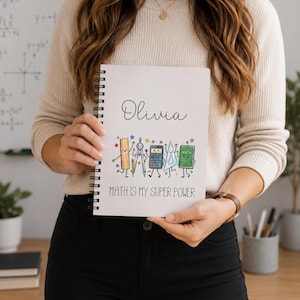 May include: A white spiral-bound notebook featuring the name Olivia in black script, followed by a colourful illustration of cartoon maths supplies including a ruler, compass, calculator, triangle, and book, with the text MATH IS MY SUPER POWER.