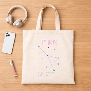 May include: Cream-colored tote bag with the word "TAURUS" in pink script, a constellation design, and the name "Olivia" with the date "20th of May". The bag is on a wooden surface, with headphones, a phone, and lip gloss.