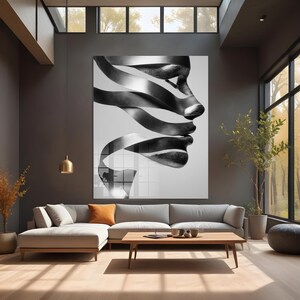 May include: Large black and white wall art featuring a profile of a face composed of flowing ribbons. The artwork hangs above a gray sectional sofa in a modern living room with a wooden coffee table and large windows.