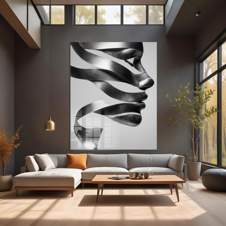 May include: Large black and white wall art featuring a profile of a face composed of flowing ribbons. The artwork hangs above a gray sectional sofa in a modern living room with a wooden coffee table and large windows.