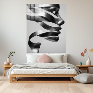 May include: Black and white abstract art print of a human profile. The artwork features a face with ribbon-like elements forming the lips and nose. The print is displayed above a bed with a wooden frame, pillows, and a blanket.