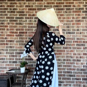 May include: A woman wearing a black and white floral dress with a white underskirt. She is wearing a traditional Vietnamese conical hat and is standing in front of a brick wall.