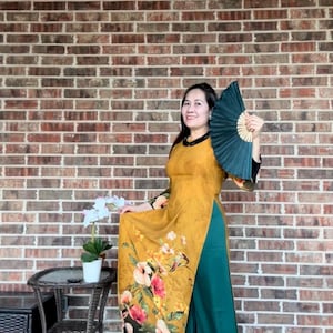 May include: A woman wearing a yellow and green dress with floral print and a green hand fan. She is standing in front of a brick wall.