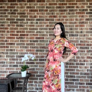 May include: A woman wearing a long, pink and orange floral print dress with a white panel down the side. She is standing in front of a brick wall.