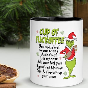 May include: White ceramic mug with a black interior featuring a Grinch character in a Santa hat and the text "CUP OF FUCKOFFEE". The mug has a humorous message with snowflakes.