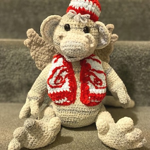 May include: A handmade crochet dragon doll in cream, gray, and red. The dragon wears a red and white striped hat and scarf. The dragon has wings and is sitting on a gray surface.