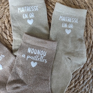 May include: Close-up of several pairs of gold-colored socks. Two pairs have the text "MAITRESSE EN OR" and a heart, while another pair says "NOUNOU a paillettes" with a heart. The socks are on a woven, light brown surface.