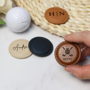 Könnte beinhalten: Vier runde Leder-Golfballmarker. Ein Marker ist braun mit den Initialen "H|N" graviert. Ein weiterer Marker ist braun mit dem Text "NETTS" und einem Design aus Golfschläger und Ball. Ein dritter Marker ist beige mit dem Namen "Andre" graviert. Der letzte Marker ist schwarz.