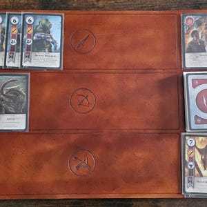 Könnte beinhalten: Ein braunes Leder-Spielbrett mit Kartenfächern, das Fantasy-Karten enthält. Die Karten zeigen Illustrationen von Kreaturen und Charakteren mit Zahlenwerten und Symbolen. Das Brett hat geprägte Symbole eines Schwertes, eines Bogens und Pfeils sowie gekreuzter Pfeile.