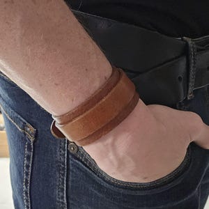 Könnte beinhalten: Ein braunes Lederarmband, das an einem Handgelenk getragen wird. Das Armband besteht aus zwei Lagen Leder und hat ein schlichtes Design.