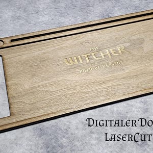 Puede incluir: Organizador de escritorio de madera con el texto "THE WITCHER PATH OF DESTINY" grabado en la superficie. El organizador tiene forma rectangular con un recorte y varios agujeros circulares. También es visible el texto "DIGITALER DOWNLOAD LASERCUT 3MM".