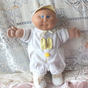May include: A doll with a round face, blue eyes, and a soft body wearing a white velour outfit with a yellow gingham bunny ear applique on the front, a matching yellow gingham cap, and white shoes with laces tied in a bow at the front.