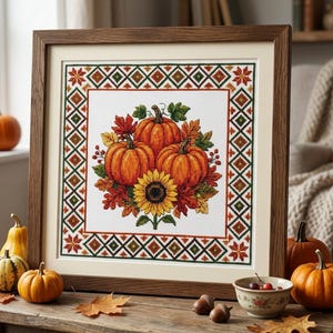 Könnte beinhalten: Ein gerahmtes Kreuzstich-Stickbild mit drei orangefarbenen Kürbissen, einer großen gelben Sonnenblume und Herbstblättern, umgeben von einer geometrischen Bordüre aus Rauten und Sternen in Grün-, Rot- und Orangetönen auf weißem Stoff.