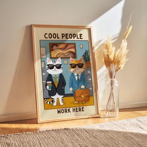May include: A framed art print showing two cartoon cats in suits and sunglasses. One cat holds a coffee cup, the other a briefcase. The print reads "COOL PEOPLE WORK HERE". The artwork is in a wooden frame and is set against a neutral background.