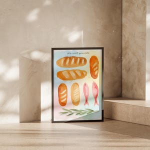 May include: A framed watercolour print depicting several loaves of bread and two pink fish. The text "the wild provide" is at the top. The artwork is in a black frame, set against a neutral background.