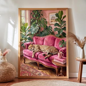 Peut inclure: Une peinture encadrée représentant un guépard dormant sur un canapé rose vif. La scène comprend des plantes vertes luxuriantes, une petite table d'appoint avec une tasse rose et deux livres empilés intitulés Wilderness Dreams et Spots & Shadows.