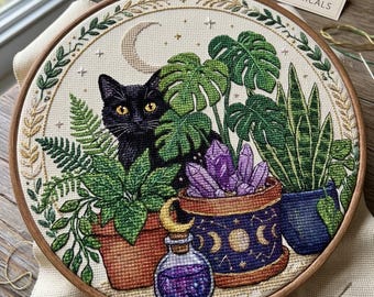 May include: A circular embroidery hoop containing a cross-stitch design of a black cat nestled among potted plants, including a monstera and snake plant. A purple crystal cluster, a small potion bottle, and a crescent moon are also stitched.