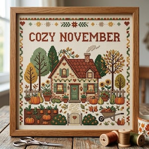 Puede incluir: Un bordado en punto de cruz enmarcado que muestra una casa con techo rojo, rodeada de árboles, calabazas y un jardín. El texto COZY NOVEMBER está bordado en la parte superior. El diseño incluye una valla, una carretilla y una puerta en tonos otoñales.